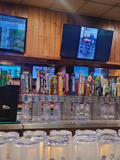 A photograph shows a bar counter from the perspective of someone standing in front of it, with various beer taps lined up along the bar. Brands include Stella Artois, Kona Big Wave, Heineken, Modelo, Peroni, Elysian Space Dust IPA, Yuengling, Michelob Ultra, Bud Light, Budweiser, and Mango Cart. Clear glass beer mugs are stacked next to the taps.

Multiple televisions are visible on the wall behind the bar. One screen shows a person lifting weights. Another shows text reading "LITY CAN BE IN OUTDOORS" and "INDOOR AIR QUALITY."

Based on the presence of brands and general aesthetics, the location is likely a bar in Kendall, United States of America.