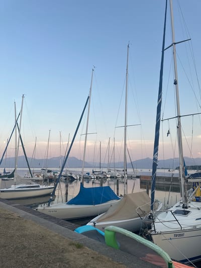 The image depicts a serene marina in Gstadt am Chiemsee, Germany, at dusk. Numerous sailboats are moored, their masts reaching towards a pale blue and pink sky. The water of Chiemsee is calm, reflecting the soft twilight hues and the silhouettes of the distant mountains. A wooden dock runs along the foreground, with a few boats tied to it. Some sailboats are covered with tarps, while others are partially visible. The atmosphere is peaceful and quiet, with the gentle light of the setting sun casting a tranquil glow over the scene.