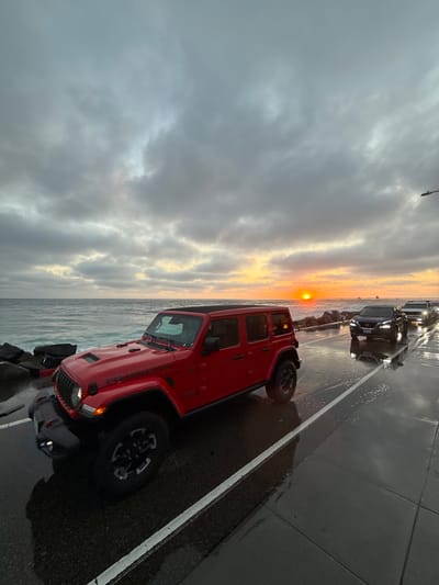 A red Jeep Wrangler Rubicon is parked on the side of a wet road next to the ocean during a cloudy sunset. The sun is setting over the Pacific Ocean, casting an orange glow on the horizon. The road is reflective due to recent rain or mist, showing the reflections of the cars and the sky. Several other vehicles are parked or driving on the road, and a pier can be seen in the distance. The scene appears to be along the coast of Oceanside, California, USA.
