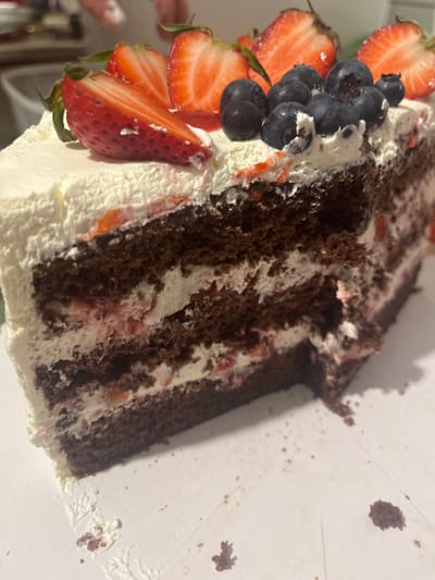 A slice of layered chocolate cake rests on a white surface, possibly a plate or countertop. The cake is decorated with fresh strawberries and blueberries along the front edge. The slice reveals multiple layers of chocolate cake alternating with white frosting and a strawberry filling. The environment is nondescript, with only a glimpse of what appears to be a kitchen setting in the background. There is no indication of the location being Rudartsi, Bulgaria, or any other specific city. No people are visible.