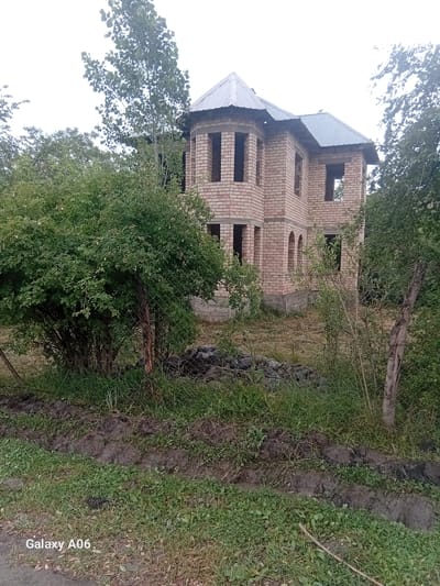 The image shows a two-story brick house under construction in Kojo-Aryk, Kyrgyzstan. The house has a metal roof and several unfinished window openings. A large, leafy bush is in the foreground, partially obscuring a wire fence. Trees and overgrown vegetation surround the property. The ground is uneven with patches of grass, mud, and what appears to be dry straw. The sky is overcast. The bottom left corner of the image displays the text "Galaxy A06".
