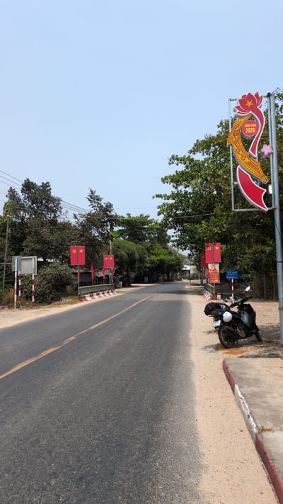 The scene depicts a street in Lâm Sơn, Vietnam. A black motorcycle is parked on the right side of the road, next to a sidewalk with a red and white curb. The road is paved with asphalt and has a yellow line marking the center. Along the left side of the road, there are several red flags with a hammer and sickle emblem. A large sign with Vietnamese script and decorative elements is also visible on the left. Trees line both sides of the street, and the sky is clear and blue.