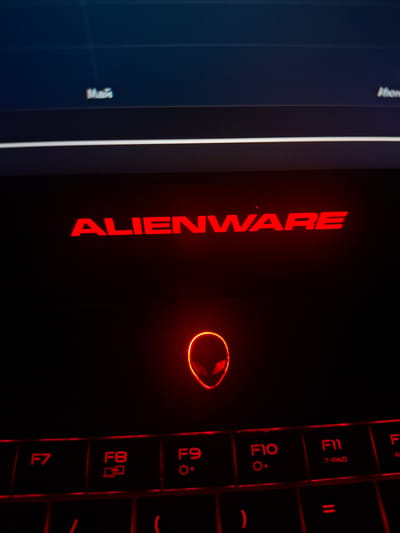 The image shows a close-up of an Alienware laptop with red illuminated accents. The Alienware logo is prominently displayed in red on the lid. Below the logo, the Alienware's iconic alien head symbol is also illuminated in red, situated above the keyboard. The keyboard itself is black with red backlighting, and several function keys (F7, F8, F9, F10, F11) are visible. The overall setting appears to be indoors, with dim lighting contributing to the glowing effect of the laptop's features. There is no visible indication of the city of Užuežerė, Lithuania, within the image.