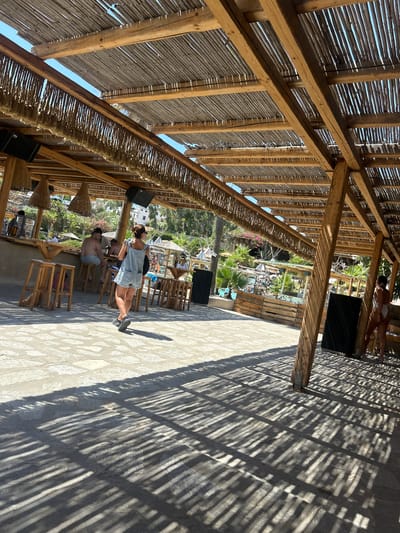 The image depicts a sun-drenched outdoor establishment in Piso Livadi, Greece, likely a beach bar or restaurant. The scene is characterized by a rustic, open-air structure with a thatched roof supported by wooden beams, casting linear shadows on the stone-paved ground. In the foreground, a woman wearing a denim romper is walking away from the camera. To the left, a bar area is visible with wooden stools and patrons seated. In the background, glimpses of a swimming pool, lounge chairs, and lush vegetation suggest a resort or vacation setting. The overall atmosphere is relaxed and summery.