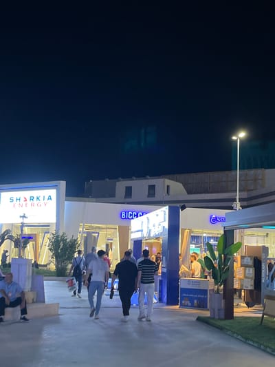 The image depicts an outdoor scene at night, likely at an event or exhibition. In the foreground, several people are walking away from the camera on a paved walkway. To the left, a large illuminated sign displays "SHARKIA ENERGY" in white and pink lettering. Further into the scene, there are architectural elements and more illuminated signs, including one that reads "BICCA" in blue neon. A street lamp is visible on the right side of the image, illuminating the area. The background shows building structures and a dark sky. The location is identified as Al-Ghizlaniya, Syria.