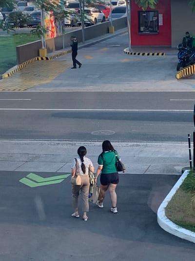 In Angeles City, Philippines, two women are walking away from the camera on a paved surface. The woman on the left is wearing a patterned blouse and tan pants, carrying a tan shoulder bag. The woman on the right is wearing a green shirt and black shorts, carrying a black shoulder bag and a shopping bag. They are walking towards a street with a green arrow painted on the pavement. In the background, a red building with a drive-thru window is visible, with a motorcycle parked nearby. A person in dark clothing is walking on a raised pathway in the background, with several cars parked beyond it. The environment appears to be a commercial area.