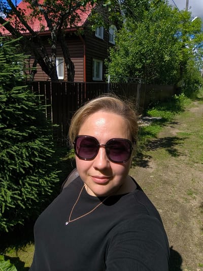 A woman is taking a selfie outdoors, with a wooden house and a fence in the background. She is wearing sunglasses and a black shirt with a gold chain and a small pendant. A large green conifer tree is to her left. A dirt path runs along the right side of the image, bordered by grass and greenery. The sky is visible with some clouds. The scene appears to be in a rural or suburban setting, possibly in Novonikolskoye, Russia.