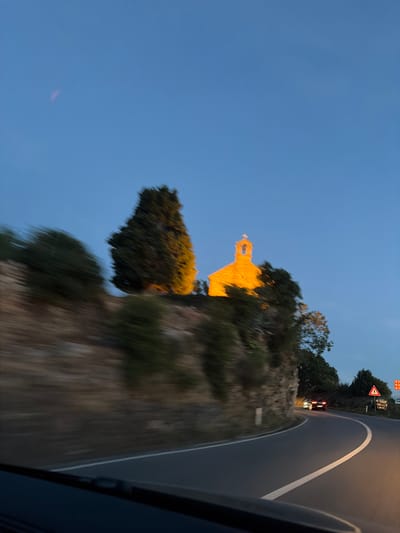 The image depicts a winding road at dusk, likely in or near Blizikuće, Montenegro. A small, stone building, appearing to be a church, is illuminated by artificial lights on a hill overlooking the road. The sky is a deep blue, transitioning from daylight to night. The road curves to the right, with a white line marking its edge. Two cars are visible on the road, one in the distance with its brake lights illuminated. The surrounding landscape is characterized by a rocky slope and trees, some of which are blurred due to motion. Road signs are also present, indicating potential hazards or directions.