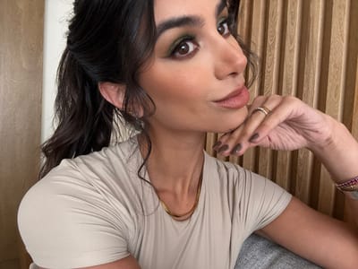 The image presents a close-up, head-and-shoulders portrait of a female subject, centrally framed. The subject has dark, wavy hair styled in a half-up ponytail. Her facial features include defined eyebrows and prominent eye makeup, specifically a dark green eyeshadow, with her eyes looking towards the camera. Her lips are coated with a light, natural-toned lipstick. She wears a plain, light beige short-sleeved top and a thin, gold-colored chain necklace. Her right hand is positioned with fingers under her chin, displaying fingernails painted in a dark olive or brown shade. A silver ring with a golden band insert is visible on her right ring finger. On her left wrist, partially visible, are a wide silver bracelet adorned with what appear to be small red stones and a narrower, plain silver bracelet. The background consists of vertical light-colored wooden slats on the right side of the frame and a plain light wall section on the left. A textured gray surface, possibly a cushion or sofa, is visible in the lower right foreground. The described location is San Luis Potosí City, Mexico.