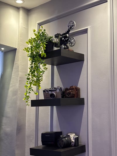 The image depicts an interior scene, likely a home or shop, in Sweileh, Jordan, featuring a collection of decorative items displayed on black floating shelves mounted on a pale grey, paneled wall.
On the top shelf, a vintage-style film projector is positioned, accompanied by a potted plant with cascading green foliage. The plant's leaves are a vibrant green, and some appear to be illuminated by ambient light.
The middle shelf displays two camera-themed objects. On the left, a metallic camera with a brown finish is visible. To its right is a wooden model of a camera, showcasing intricate detailing.
The bottom shelf holds two more cameras. On the left, a black Polaroid Autofocus 660 camera is present. To its right, a silver and black digital camera with a lens is positioned.
The environment appears to be dimly lit, with shadows cast on the wall by the shelves and objects. The wall itself has a textured finish and is divided by vertical molding. Two recessed ceiling lights are partially visible at the top of the frame, suggesting the primary light source. No people are present in the scene.