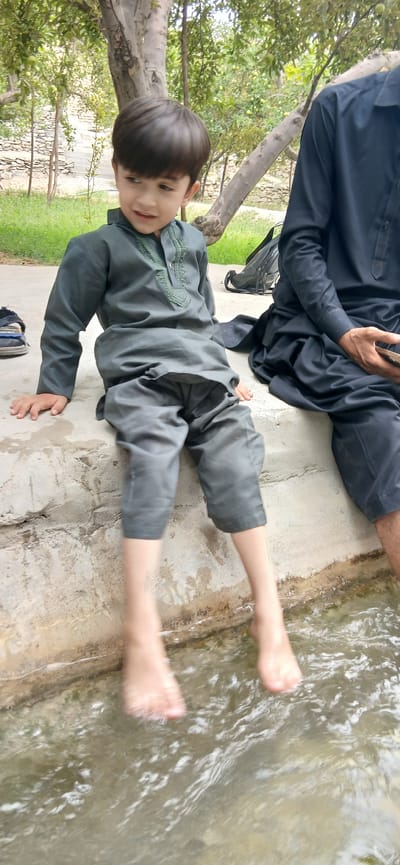 In Kan Mehtarzai, Pakistan, two individuals, a young boy and an adult male, are seated on a concrete ledge next to a body of water. The boy, wearing a grey shalwar kameez, is positioned on the left, with his feet submerged in the water. His gaze is directed to his left, and his mouth is slightly open, suggesting he is speaking or smiling. The adult male, on the right, is partially visible, wearing a dark shalwar kameez and holding a mobile phone. A black bag rests behind the boy. The background features trees and a stone wall, indicating an outdoor, possibly natural, setting within the city.