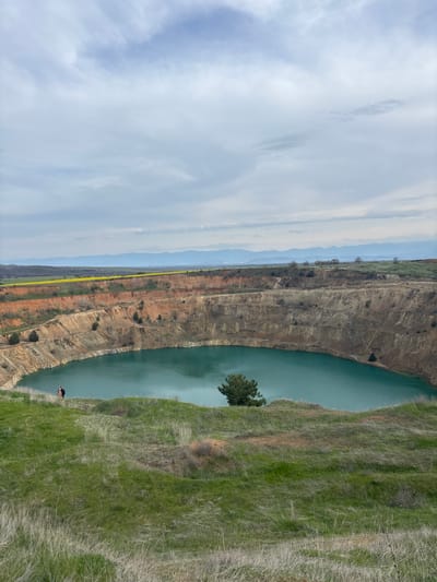 The image captures a portion of the "Emerald Lake" quarry in Tsar Asen, Bulgaria. The quarry pit, filled with turquoise water, occupies the center of the frame, with a tree growing near the water's edge. The quarry walls display layered earth tones, indicating the depth and composition of the excavated area. A strip of vegetation and distant mountains can be seen in the background. On the right, the grassy bank slopes down to the quarry's edge. A person in dark clothing stands on the quarry edge. The sky is overcast with a mix of clouds.