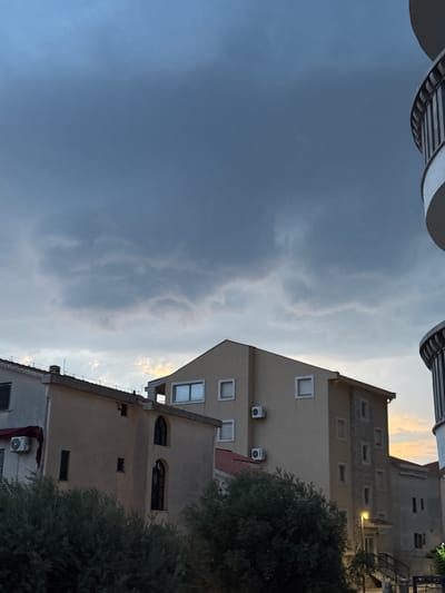 The image depicts a view looking upwards at buildings and the sky. The perspective is from a low angle, possibly from ground level or a lower floor of a building.
In the foreground, a portion of a balcony railing with a striped pattern is visible at the top. The majority of the image is dominated by a cloudy sky, with some patches of lighter clouds suggesting either an approaching storm or the aftermath of one, with hints of warm, sunset-like light peeking through.
The background features several buildings, appearing to be residential or mixed-use. These buildings are of varying heights and architectural styles. One prominent building is a lighter beige color and has several rectangular windows, some with visible shutters or frames. Air conditioning units are mounted on the exterior walls of this building. Another building is darker, with smaller, rectangular windows. The rooftops of some buildings are visible, with one showing a red tiled section and another with what appears to be a chimney.
The overall scene suggests an urban or semi-urban environment. The time of day appears to be late afternoon or early evening, given the lighting conditions in the sky. The weather appears to be overcast with dramatic cloud formations. There is no visible text in the image. The atmosphere conveyed is somewhat dramatic due to the sky, but also mundane due to the focus on buildings.