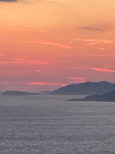 This image depicts a serene seascape at sunset, with a warm orange and pink sky illuminating the horizon. The sky transitions from a pale yellow at the top to a vibrant pink and orange, streaked with wispy clouds. Below the sky, a range of dark, silhouetted mountains recedes into the distance, their forms softened by the atmospheric haze. The foreground is dominated by a calm body of water, reflecting the muted colors of the sky. The water's surface is textured with subtle ripples, indicating a gentle breeze. In the middle ground, several islands or peninsulas are visible as dark shapes against the water. The image captures the tranquil beauty of a coastal landscape, likely in Glavatičići, Montenegro, known for its picturesque Adriatic scenery. There are no people or explicit actions depicted, focusing solely on the natural environment.