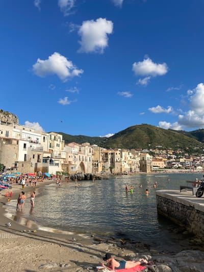 The image displays a coastal scene in Cefalù, Italy, specifically focusing on the Spiaggia della Marina (Cefalù Beach) with the historic town and its mountainous backdrop.
The environment is characterized by a bright blue sky with several distinct white cumulus clouds. A dark, small bird is visible in flight in the upper-left quadrant. Green, treed mountains rise in the background, with urban development extending partway up their slopes. The foreground and midground feature a sandy beach transitioning into a calm, dark blue-green bay. To the right, a stone pier or wall extends into the water, on which a dark motorcycle is parked next to a wooden bench. Buildings of varying heights, predominantly light-colored (cream, beige, off-white), line the waterfront. These structures feature multiple stories, windows, and balconies. A notable architectural element, the Porta Pescara (Fisherman's Gate arch), spans a rocky section where buildings meet the water on the mid-left. Shadows cast by buildings and people suggest strong sunlight, indicating daytime.
Numerous people are present across the scene, engaged in leisure activities. On the sandy beach, individuals are standing, walking, and sitting, with some under colorful beach umbrellas (red, blue, striped). In the bay, a considerable number of people are swimming, wading, or standing in the water, distributed throughout the visible area. One individual in the immediate foreground is lying prone on a red towel on rocks, wearing dark swimwear and a dark hat, appearing to read a book or device. Most visible individuals are wearing swimwear. Objects include beach towels, umbrellas, and what appear to be small beach toys. The scene depicts a lively, populated beach and coastal area.