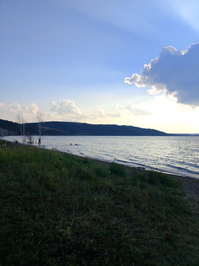 A wide shot captures a serene lake scene under a partly cloudy sky with sunlight streaming through gaps in the clouds. The foreground is dominated by a grassy slope leading down to a sandy shore. The lake stretches to the horizon, with a tree-covered hill forming the backdrop on the far side. Several bare trees stand on the left side of the shore, and a few people are visible in the shallow water, swimming or wading. The water is calm with gentle ripples. Birds are scattered across the sky. The location is identified as the city of Mnogosopochnoye, Kazakhstan.