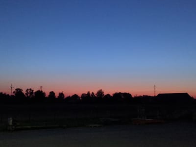 The image depicts a twilight landscape in Bolesławów, Poland. The sky transitions from a deep blue at the top to a soft pink and orange hue near the horizon, indicating either dawn or dusk. A silhouetted line of trees forms the dominant feature against the colorful sky, with the dark shapes of deciduous trees and some evergreens.
To the left of the image, a wind turbine stands tall, its blades visible against the sky. Further to the right, a power line pylon with crossbars is also silhouetted. In the far right of the frame, a dark, angular structure, possibly a barn or outbuilding, is visible in silhouette.
The foreground is a dark, unlit area that appears to be a rural or semi-rural setting, possibly a yard or field. Some indistinct objects, like pallets or crates, are faintly visible in the mid-ground, suggesting human activity or storage. The ground in the immediate foreground is a dark, paved surface, possibly a road or driveway. The overall impression is one of a quiet, rural evening or morning in the Polish countryside.