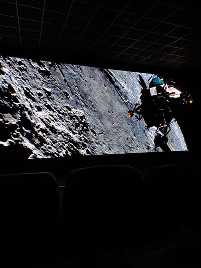 The image captures a forensic view inside an immersive theater or planetarium, specifically the Space Adventure Canela in Canela, Brazil. The dominant feature is a wide, large projection screen displaying a detailed, monochrome depiction of the heavily cratered lunar surface, which occupies the upper two-thirds of the frame. In the upper right quadrant of the screen, the silhouette of a Lunar Module (LM) is visible against the lunar backdrop, its complex structure, landing gear, and antennae clearly discernible. Faint, localized orange-yellow glows are present near the ends of some of the LM's protrusions, possibly indicating thruster activity or reflective surfaces. The room is almost entirely dark, with illumination provided solely by the screen's projection. In the lower third of the image, the dark, featureless silhouettes of two upholstered theater-style seats are visible from a rear perspective, positioned for viewing the screen. The ceiling above is dark with a distinct grid pattern, characteristic of acoustic ceiling tiles in a public venue. No individuals are present or visible within the scene. The overall environment suggests an immersive educational or entertainment experience focused on space exploration.