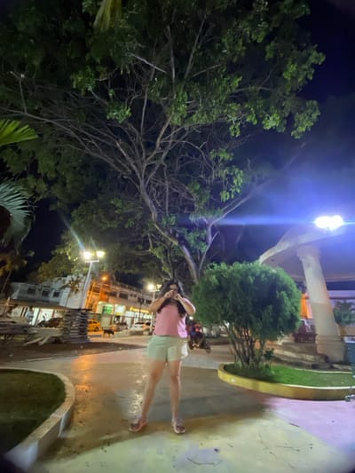 The image depicts a nighttime scene in a public park or plaza within the city of General Villamil Playas, Ecuador. A lone individual, appearing female, stands centrally, facing the camera. She wears a light pink short-sleeved top, light green shorts, and pink sandals. Her dark hair is long, and her hands are raised to her face, partially obscuring it.
Dominating the upper frame is a large tree with extensive branches and green foliage, silhouetted against the dark night sky. The ground consists of a paved surface with curved concrete borders defining grassy areas. To the right, a white, columned gazebo-like structure is partially visible, adjacent to a dense green shrub.
In the background, to the left of the main subject, are multi-story buildings with illuminated windows and storefronts, indicating an urban street. Several vehicles, including cars and a yellow taxi, are parked or moving on the street. A dark-colored dog is visible near the vehicles. Streetlights with bright, warm yellow illumination cast light on the background buildings and street. A very bright, blue-white light source, likely a streetlamp, is present in the upper-right quadrant, causing a prominent lens flare. To the far left, a stack of dark, rectangular objects, possibly construction materials, is visible. The overall scene is artificially lit, suggesting a lively public area after dark.