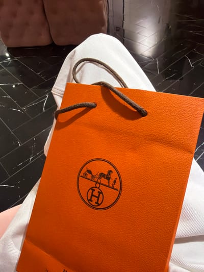 An orange Hermes shopping bag with a brown rope handle is held in the foreground, resting on the lap of a person wearing white pants. The bag features the iconic Hermes logo, a horse-drawn carriage with a charioteer and the letter "H" centered within a circle. The background consists of a dark, patterned floor and the blurred outline of a pink upholstered chair. The scene appears to be indoors, likely a retail environment, and there is no specific indication of the location within Xuanwu District, China, other than the provided context.