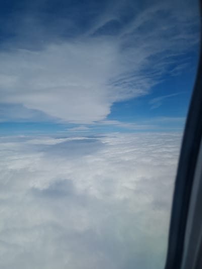 The image shows a view of the sky from an airplane window. The top of the window frame is visible. The sky is predominantly blue with varying shades and cloud formations. There is a distinct horizon line separating the sky from what could be either more clouds or the earth's surface in the distance. The location cannot be determined as Breze, Bulgaria or any other specific place based on the image.