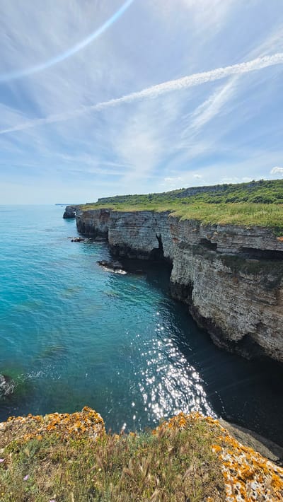 The image captures a coastal scene at Kamen Bryag, Bulgaria. A layered cliff face, topped with green vegetation, borders the turquoise water of the Black Sea. Small caves are visible in the cliff. The sea reflects sunlight, creating bright patches. A cliff with yellow lichen and grass appears on the right side. The sky is blue with some contrails.