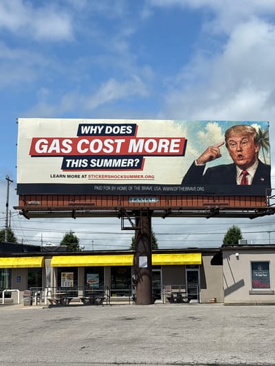 A large billboard is prominently displayed against a cloudy blue sky, featuring a portrait of Donald Trump. The billboard's text reads: "WHY DOES GAS COST MORE THIS SUMMER? LEARN MORE AT STICKERSHOCK SUMMER.ORG PAID FOR BY HOME OF THE BRAVE USA. WWW.OFTHEBRAVE.ORG". Below the billboard, a brick-colored structure supports it, with a smaller sign that reads "LAMAR".
In the foreground, a parking lot with cracked asphalt stretches out. Behind the parking lot is a low-lying building with yellow awnings over its windows and doors. The building appears to be a commercial establishment, possibly a restaurant, with picnic tables outside. Signs on the building indicate it is open. Power lines and utility poles are visible in the background, as well as trees and other buildings. The overall scene suggests an urban or suburban environment in Columbus, United States of America.