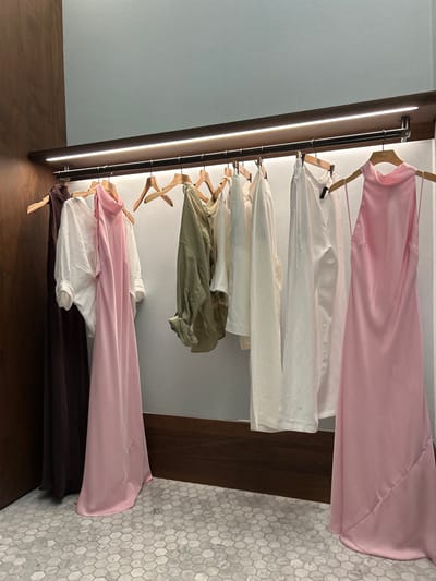 This is a vertically oriented, eye-level medium shot of a clothing rack with several garments hanging on wooden hangers. The rack is attached to a dark wooden structure that runs vertically along the left side of the frame and appears to be a closet or a wall unit. The clothing items are predominantly in shades of pink, white, and olive green, with a dark brown item at the bottom.
The garment at the very top is a light pink, possibly satin, dress or top. Below it are several white garments, some appearing to be shirts or blouses. Further down is an olive green garment, possibly a shirt or jacket, followed by another white garment. The lowest visible garment is a dark brown piece of clothing. The garments are neatly arranged, suggesting an organized closet or display.
The background on the right consists of a hexagonal mosaic tile pattern in shades of white and gray, which transitions into a dark wooden surface. The background on the left is a plain, light blue wall, with a thin, bright vertical light source illuminating the wooden structure. The lighting suggests an indoor setting, possibly a home closet, boutique, or retail space. There is no visible activity or people in the image. The overall mood is calm and orderly. No visible text is present in the image. The location context is Aventura, United States of America, but this context is not directly inferable from the visual content.