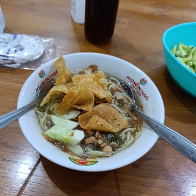 A bowl of noodles with fried wonton chips, cucumber slices, and a brown broth is on a wooden table. A metal spoon is laying on top of the noodles. The bowl says "WOWIN Food" in black lettering on a red background. The table appears to be in a food stall or restaurant. The location is in Kecamatan Asembagus, Indonesia, at coordinates -7.75 latitude and 114.21 longitude. The image shows no people.