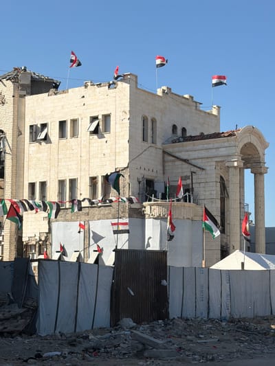 The image shows a damaged building in Jabalia, Palestinian Territories. The building, constructed of light-colored stone blocks, has several flags flying from its upper levels and balconies. The flags appear to be Palestinian flags and Egyptian flags. The roof of the building is partially collapsed and shows signs of damage. In front of the building, there are temporary structures made of white tarpaulins and corrugated metal, possibly shelters or aid distribution points. Debris, including rubble and scattered objects, is visible on the ground in the foreground, suggesting destruction or conflict. The sky is clear and blue. The scene appears to depict a post-conflict or disaster-affected area.
