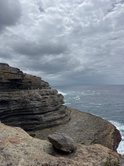 A striking coastal vista presents a dramatic interplay of textures and light. A sheer cliff face plunges towards the sea, its stratified rock layers etched by time and weather. A detached boulder clings precariously to the edge. Above, a turbulent sky is filled with swirling cloud formations. The ocean stretches to the horizon, its surface churned by wind and waves. The scene evokes a sense of natural power and rugged beauty.