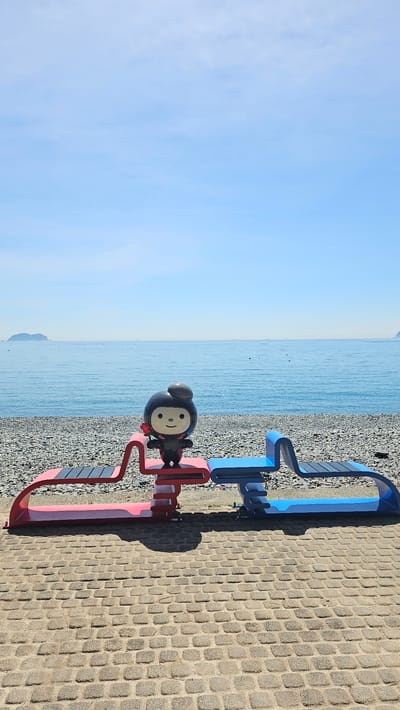 A cartoon-like mascot stands on a red and blue abstract sculpture facing a calm, blue ocean under a clear sky. The mascot is black with a round head, a smile, and a small grey hat. It holds a red flower in its right hand. The sculpture is situated on a paved walkway, with a pebbled beach and the sea stretching to the horizon. In the distance, a small island is visible on the left, and another landmass on the right. The scene is likely located in Geoje-si, South Korea, given the prevalence of coastal areas and the distinct aesthetic of public art installations in the region.