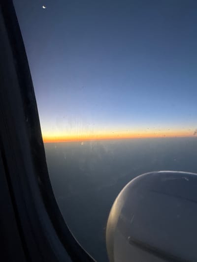 The image depicts a view from an airplane window during what appears to be either sunrise or sunset. The sky transitions from a deep blue at the top to a pale orange horizon line. A sliver of a crescent moon is visible in the upper left corner. A dark, textured curtain lines the left edge of the frame, obscuring part of the view. In the lower right corner, a portion of an airplane engine is visible. The scene is set above a layer of clouds or haze, with the landscape below indistinct. The image is grainy, suggesting it was taken in low light conditions or with a mobile device. The provided crops offer closer views of the moon, the sky, the horizon, and the airplane engine. Based on the provided information, this image is not specifically located in Monoq, Uzbekistan, as there is no information within the image or its crops to suggest this location.