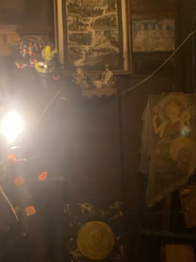 The image, potentially taken in Baan Don Sai, Thailand, shows a cluttered interior space. Hanging on a dark wall are two framed images, one above the other on the left. Further right, an indistinct plastic bag containing various items hangs from the ceiling. A decorative plate is mounted on the wall in the upper right corner. Below the plate are faint, blurry images of orange flowers. A light source obscures the bottom of the frame.