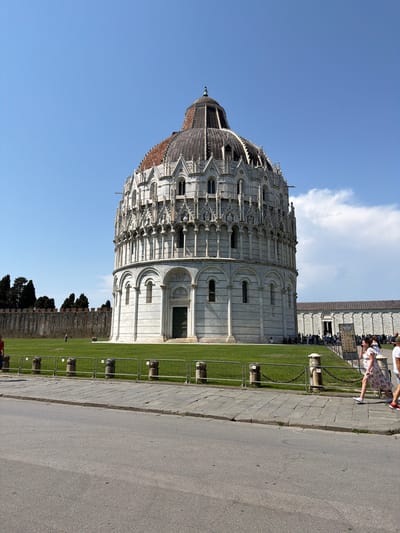 The image shows the Baptistery of Pisa, a circular building with a dome, located in Pisa, Italy. The Baptistery is made of white marble and is surrounded by a green lawn and a stone wall. There are several people walking around the Baptistery, and some are taking pictures. The sky is blue with a few white clouds.