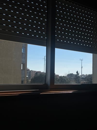 The image displays a daytime view from an interior looking out through a window onto an urban environment in Pedrouços, Portugal. The top portion of the window opening is obscured by partially lowered dark grey roller blinds, characterized by horizontal slats and small, evenly distributed white perforations. The window features dark framing and a central vertical mullion. Below the transparent glass panes, a wooden windowsill is present, with a distinct band of direct sunlight illuminating its surface.
Beyond the window, a clear, bright blue sky with a faint contrail in the upper right dominates the background. To the immediate left, a multi-story building with light brown brickwork is visible, featuring a single rectangular window. Adjacent to it, a segment of a lighter grey building is apparent. In the mid-ground, a tall, metal lattice transmission tower stands prominently, with overhead utility lines extending horizontally. Further to the right, a shorter utility pole equipped with crossarms is observed. A dense line of dark green trees occupies the middle distance, partially concealing several multi-story buildings with reddish-brown facades. No people, specific actions, or interactions are visible within the scene.