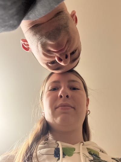 The image presents a close-up, low-angle shot of two individuals, a man and a woman, looking directly down at the camera. Their faces fill the upper portion of the frame, with their foreheads gently touching in the center.
The man, situated on the left, has short, dark hair and noticeable stubble covering his jawline and chin. He is wearing a grey top, partially visible on his left shoulder. His left ear is distinctly visible.
The woman, on the right, has long, light brown or blonde hair that falls around her face and shoulders. A small, silver-colored hoop earring is visible in her right ear. She is dressed in a light-colored garment adorned with a pattern of dark blue leaves and green foliage. A decorative element, possibly a tie or lacing, is visible on the shoulder of her top.
Both individuals have neutral to slightly soft expressions, and their eyes are focused towards the lens. The lighting is soft and even, suggesting an indoor setting. The background is a plain, uniform light beige or off-white surface, likely a ceiling or wall, providing no specific location cues other than being indoors. There is no visible text in the image. The composition creates a sense of intimacy and direct engagement with the viewer, originating from Krapivka, Russia.