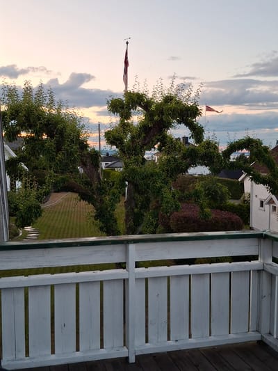 The image captures a tranquil scene from a balcony in Åsgårdstrand, Norway, overlooking a residential neighborhood during twilight. In the foreground, a weathered white wooden railing and deck frame the view. Beyond the railing, a meticulously manicured lawn with striped patterns stretches towards a curved pathway of stepping stones. A large, ornately pruned tree dominates the mid-ground, its branches extending outward and upward, creating a natural canopy. In the distance, several houses with dark roofs are nestled amongst lush greenery. Two Norwegian flags are visible, one prominently displayed on a flagpole with a weather vane, and another smaller flag fluttering gently. The sky above is painted with soft hues of pink and orange, with scattered clouds adding texture to the fading light. The overall atmosphere is one of peaceful domesticity and the beauty of a Norwegian summer evening.