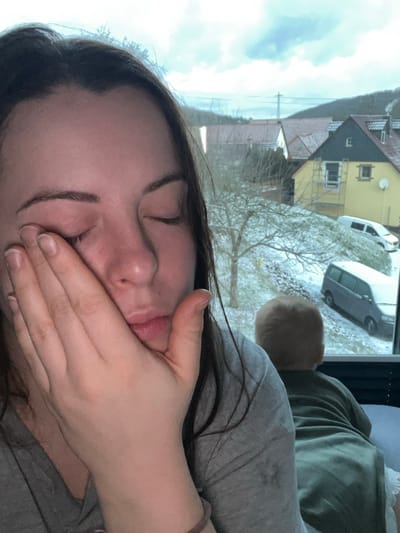 A woman is reclined, facing the camera. Her eyes are closed and a hand is covering her mouth. Behind her is a window showing a snowy scene with houses, trees, and vehicles covered in snow. A light-haired baby's head is visible between the woman and the window. The woman wears a grey shirt, and a green shirt is also visible.