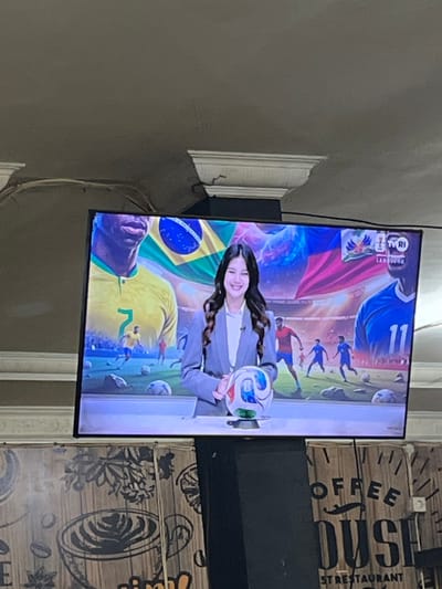 A woman is shown on a television screen, holding a soccer ball and smiling. She is wearing a gray blazer over a white shirt. The television screen displays a soccer-themed graphic with flags and players. The screen is mounted on a black pole, which is in front of a wooden wall with coffee-related drawings and text. The location is Keudesampoiniet, Indonesia.