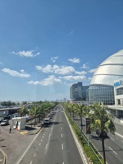 A street view in Pasay, Philippines. A multi-lane asphalt road stretches into the distance, marked with white lane dividers and bordered by palm trees. Buildings are present on the left, including one with the signage "IMAX". The sky is blue with scattered white clouds. Cars and pedestrian are present.