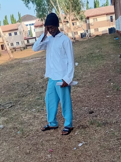 A man with a black beanie stands on a grassy field. He wears a white button-down shirt and light blue pants, with black sandals on his feet. He is holding something white in his hand and appears to be talking on a phone held to his ear. Buildings are visible in the background, and some trash litters the grass. The image is rotated to the left.