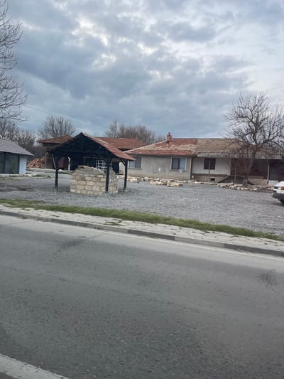 The scene depicts an outdoor environment in Osenets, Bulgaria, under an overcast sky. In the foreground, an asphalt road runs horizontally, bordered on the right by a concrete sidewalk and a thin strip of green vegetation. Beyond the sidewalk lies a large, open plot covered primarily with light grey gravel. Centrally located within this gravel area is a stone well constructed from light-colored, rectangular blocks, topped by a dark wooden canopy with a reddish-brown tiled roof. To the far right, the front bumper and headlight of a white vehicle are partially visible, parked on the gravel. Numerous irregular stones or concrete fragments are scattered across the gravel plot, particularly in front of the main building.
In the mid-background, multiple structures are visible. To the left, a smaller building with a dark roof and large window panels is present. Directly behind the well structure is a house with light-colored walls, multiple windows, and a reddish-brown tiled roof featuring a chimney. To the right of this house, another dwelling appears, distinguished by an open porch or veranda supported by dark pillars and a similar tiled roof. The background horizon features a dense line of bare deciduous trees, indicative of a dormant season. No people or active interactions are observed.