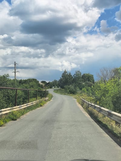 The image depicts a winding asphalt road with guardrails on either side. The road appears to be in a rural or semi-rural area, with lush green trees and vegetation lining the sides. In the distance, there are a few houses visible, suggesting a small settlement. The sky is partly cloudy, with dramatic dark clouds interspersed with patches of blue sky. A utility pole with electrical wires is visible on the left side of the road. The road itself shows signs of wear and tear, with some patches and cracks. The guardrails are made of metal and show signs of rust and minor damage. The overall impression is of a scenic but somewhat neglected rural road. The location is specified as Severnyi, Russia.
