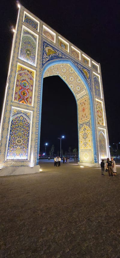 This is a nighttime, low-angle, full shot of a large, ornate archway in Satkak, Uzbekistan. The archway is intricately decorated with colorful mosaics and illuminated by white lights around the edges of the panels. The archway has a bright blue border around the opening, with geometric patterns visible within the arch and on the side panels.
In the foreground, the ground is paved with dark, textured material, possibly cobblestones or asphalt, with some scattered smaller lights at the base of the archway.
In the mid-ground, several individuals are standing and walking around the plaza. On the left side, a group of people in light-colored clothing are gathered. Further back, a few individuals are visible near streetlights. On the right side, near the base of the archway, a few more people are standing, including a child.
The background is dark, indicating nighttime, with visible streetlights and some trees suggesting an urban or park-like setting. The overall scene conveys a festive or ceremonial atmosphere.
