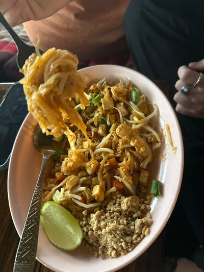 An overhead close-up image depicts a plate of Pad Thai being consumed in Ban Paem Bok, Thailand. A light pink, oval-shaped plate contains Pad Thai noodles with bean sprouts, diced tofu, chicken strips, chopped green onions, and shredded carrots. A pile of crushed peanuts is located at the bottom right of the plate, and a lime wedge rests at the bottom left. A silver-colored fork, held by an unidentifiable individual, is actively lifting a substantial portion of noodles from the top left of the plate. A silver-colored spoon with a patterned handle rests in the dish. In the background, a person wearing dark clothing has their right hand visible, featuring a silver-colored ring on the index or middle finger. A dark object, possibly a mobile phone, is partially visible on a dark wooden table surface to the left of the plate. Another piece of patterned fabric is visible on the far left.