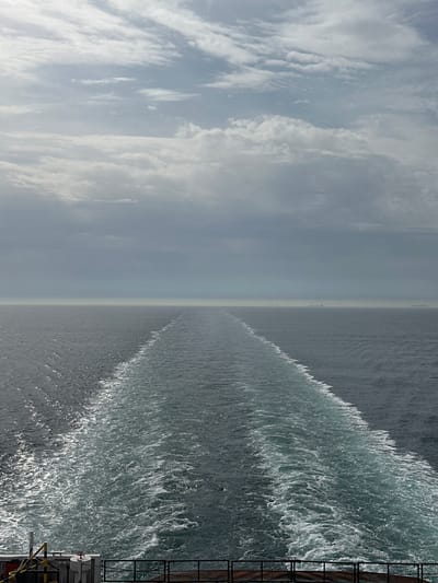 The image depicts a seascape. The sky, filled with clouds, occupies the left half, while the ocean, churning with waves, takes up the right half. The horizon line is slightly blurred due to haze. The perspective indicates the view is from a vessel, evidenced by the wake of the ship cutting through the water. A ladder and railing on a structure are visible in the lower right corner, confirming the viewer's position on a vessel. The location is an unidentified part of the sea near Unknown, France.