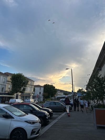 In Alès, France, on a street lined with parked cars and buildings, a person rides an electric scooter down a sidewalk. A group of individuals walks in the same direction behind the scooter rider. The sky above is partly cloudy, with patches of sunlight breaking through, suggesting either sunrise or sunset. The scene captures a moment of urban activity with a backdrop of residential and commercial architecture.