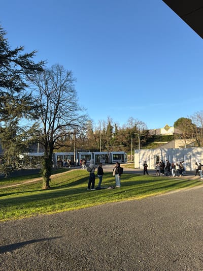 This image captures an outdoor urban landscape under a clear blue sky, indicating daylight conditions with prominent shadows suggesting late afternoon or early morning. The scene is located in Saint-Priest, France, identifiable by the "TCL" branding on the tram, a transport network serving the Lyon metropolitan area including Saint-Priest.

In the foreground, a dark grey asphalt or gravel pathway runs horizontally, exhibiting a long shadow cast towards the lower-left corner. A bright green grassy slope dominates the mid-ground. On this slope, three individuals stand in a loose group, appearing to interact. The individual on the left wears dark clothing with a light-colored hood or backpack. The central figure is dressed in dark attire, and the rightmost person wears a dark top with light-colored pants. A dark object rests on the grass near their feet.

To the left, a large coniferous tree with dark green foliage is partially visible, along with a tall, bare deciduous tree whose trunk is partially covered in ivy. Beyond these, a light grey, multi-car tram is stationary at a tram stop. Its doors are open, and multiple individuals are visible on the platform and within the tram, suggesting passenger activity.

To the right of the tram station, a low-rise, light-colored concrete building is situated. A group of approximately ten to fifteen individuals, casually dressed, are either standing or walking near this building. In the background, various trees, some bare and some with foliage, delineate the terrain, and additional buildings are visible, including one with a distinctive yellow diamond-shaped architectural feature on its roof.