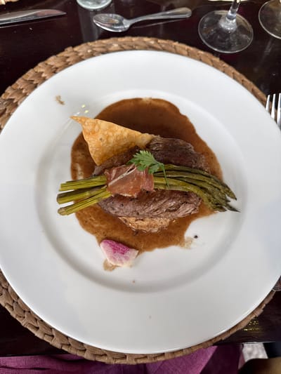 A top-down shot reveals a meal served on a white plate placed on a woven placemat. The food appears to consist of a cut of meat, possibly steak, surrounded by a brown sauce or gravy. A bundle of asparagus spears rests atop the meat, with a thin slice of prosciutto and a sprig of cilantro adorning the arrangement. A crisp, triangular chip sits adjacent to the meat, and a small slice of what appears to be a radish sits on the opposite side of the plate. Silverware and glass stemware are partially visible alongside the plate, suggesting a dining setting. The presence of a purplish textile near the edge suggests a table setting. The specific location of the city of Santiago de Querétaro, Mexico is not determined in this photo.